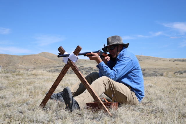 Seated cross-sticks position with a single-shot rifle on open prairie.